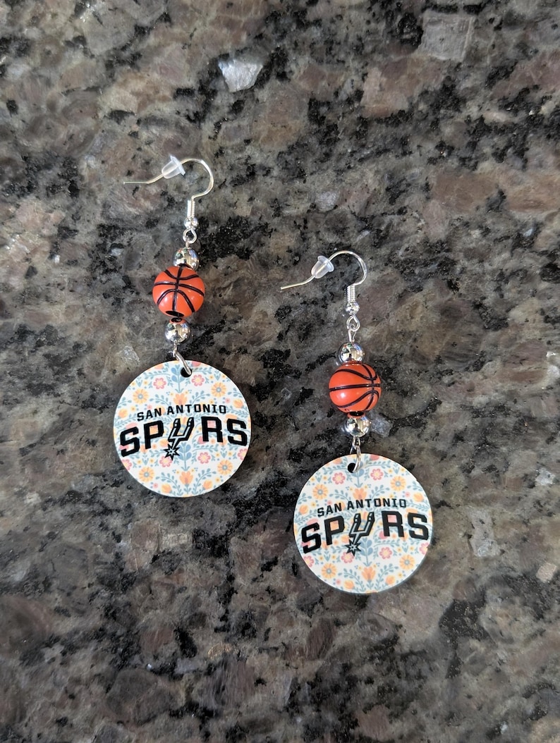 May include: A pair of dangle earrings featuring silver-toned hooks, small orange basketball beads, and circular charms. The charms display a floral pattern in soft colors with the text SAN ANTONIO SPURS printed in black, including the team logo.