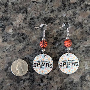 May include: A pair of dangle earrings featuring a round charm with a floral pattern and the text 'SAN ANTONIO SPURS', accented by a small orange basketball bead and silver-tone hardware, shown next to a US quarter for scale reference.