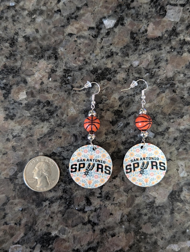 May include: A pair of dangle earrings featuring a round charm with a floral pattern and the text 'SAN ANTONIO SPURS', accented by a small orange basketball bead and silver-tone hardware, shown next to a US quarter for scale reference.