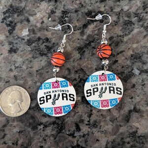 May include: A pair of dangle earrings featuring a round white charm with the text San Antonio Spurs and a basketball graphic, accented by orange basketball beads and silver-tone hardware, displayed next to a United States quarter coin for scale.