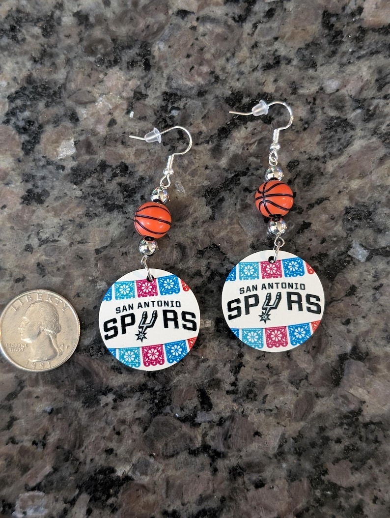 May include: A pair of dangle earrings featuring a round white charm with the text San Antonio Spurs and a basketball graphic, accented by orange basketball beads and silver-tone hardware, displayed next to a United States quarter coin for scale.