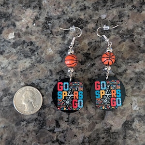 May include: A pair of dangle earrings featuring orange basketball-shaped beads and circular black pendants with the text GO SPURS GO in blue, red, and white, alongside a silver-colored US quarter coin for scale on a dark granite surface.