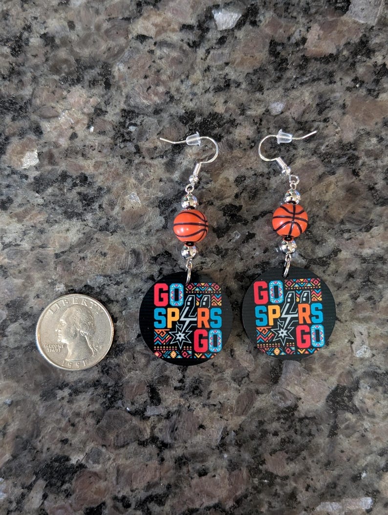May include: A pair of dangle earrings featuring orange basketball-shaped beads and circular black pendants with the text GO SPURS GO in blue, red, and white, alongside a silver-colored US quarter coin for scale on a dark granite surface.