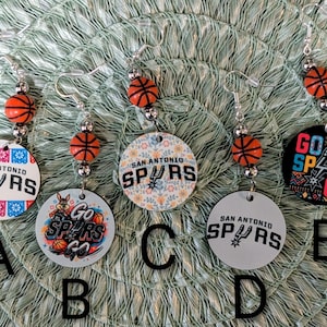 May include: Five pairs of dangle earrings featuring San Antonio Spurs basketball themes. Each earring includes a small orange basketball bead and a circular pendant with various designs, including team logos and the text Go Spurs Go, labeled A through E.