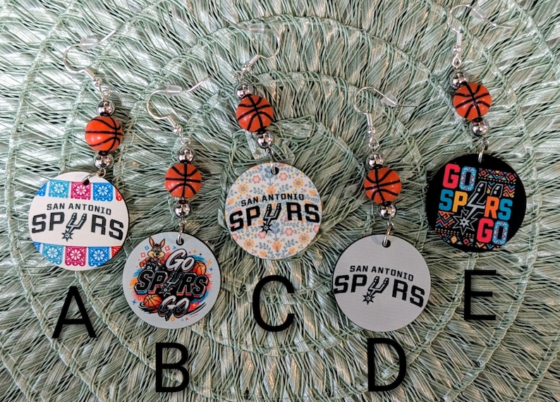 May include: Five pairs of dangle earrings featuring San Antonio Spurs basketball themes. Each earring includes a small orange basketball bead and a circular pendant with various designs, including team logos and the text Go Spurs Go, labeled A through E.