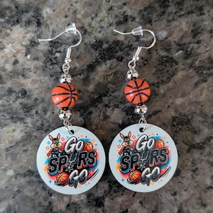 May include: A pair of dangle earrings featuring silver-toned fishhook wires, small metallic beads, and orange basketball-shaped beads. Each earring includes a circular white pendant printed with a cartoon coyote, basketballs, and the text Go Spurs Go.