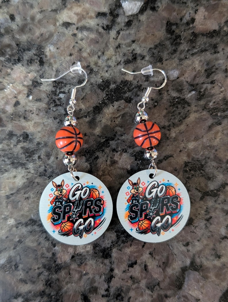 May include: A pair of dangle earrings featuring silver-toned fishhook wires, small metallic beads, and orange basketball-shaped beads. Each earring includes a circular white pendant printed with a cartoon coyote, basketballs, and the text Go Spurs Go.