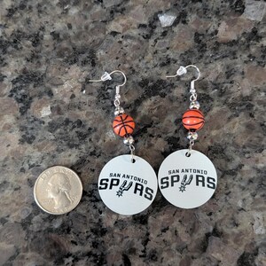May include: A pair of dangle earrings featuring a round white disc with the black text SAN ANTONIO SPURS and a team logo. Above the disc is a small orange basketball bead with black lines, connected by silver-toned metal hardware and hooks.