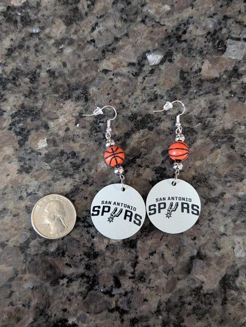 May include: A pair of dangle earrings featuring a round white disc with the black text SAN ANTONIO SPURS and a team logo. Above the disc is a small orange basketball bead with black lines, connected by silver-toned metal hardware and hooks.