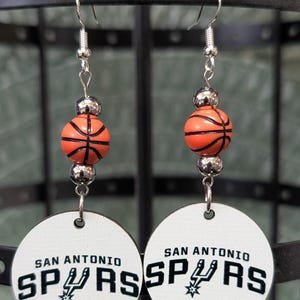 May include: A pair of dangle earrings featuring a round white disc with the black text San Antonio Spurs and a team logo. Above the disc is an orange basketball bead flanked by two small metallic silver beads on a silver-toned metal hook wire.
