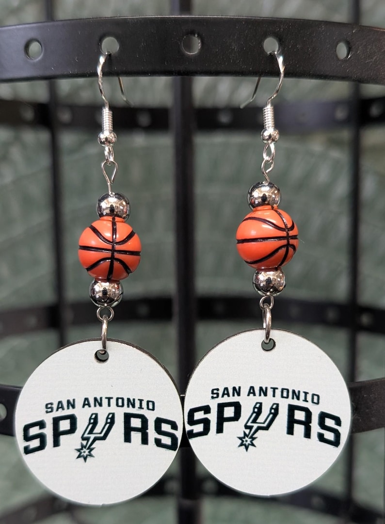 May include: A pair of dangle earrings featuring a round white disc with the black text San Antonio Spurs and a team logo. Above the disc is an orange basketball bead flanked by two small metallic silver beads on a silver-toned metal hook wire.