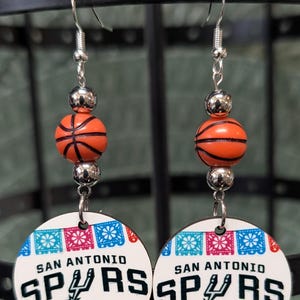 May include: A pair of dangle earrings featuring a silver-tone hook, a metallic silver bead, an orange basketball bead, and a circular white pendant. The pendant displays the text SAN ANTONIO SPURS with a team logo and colorful floral banner patterns.