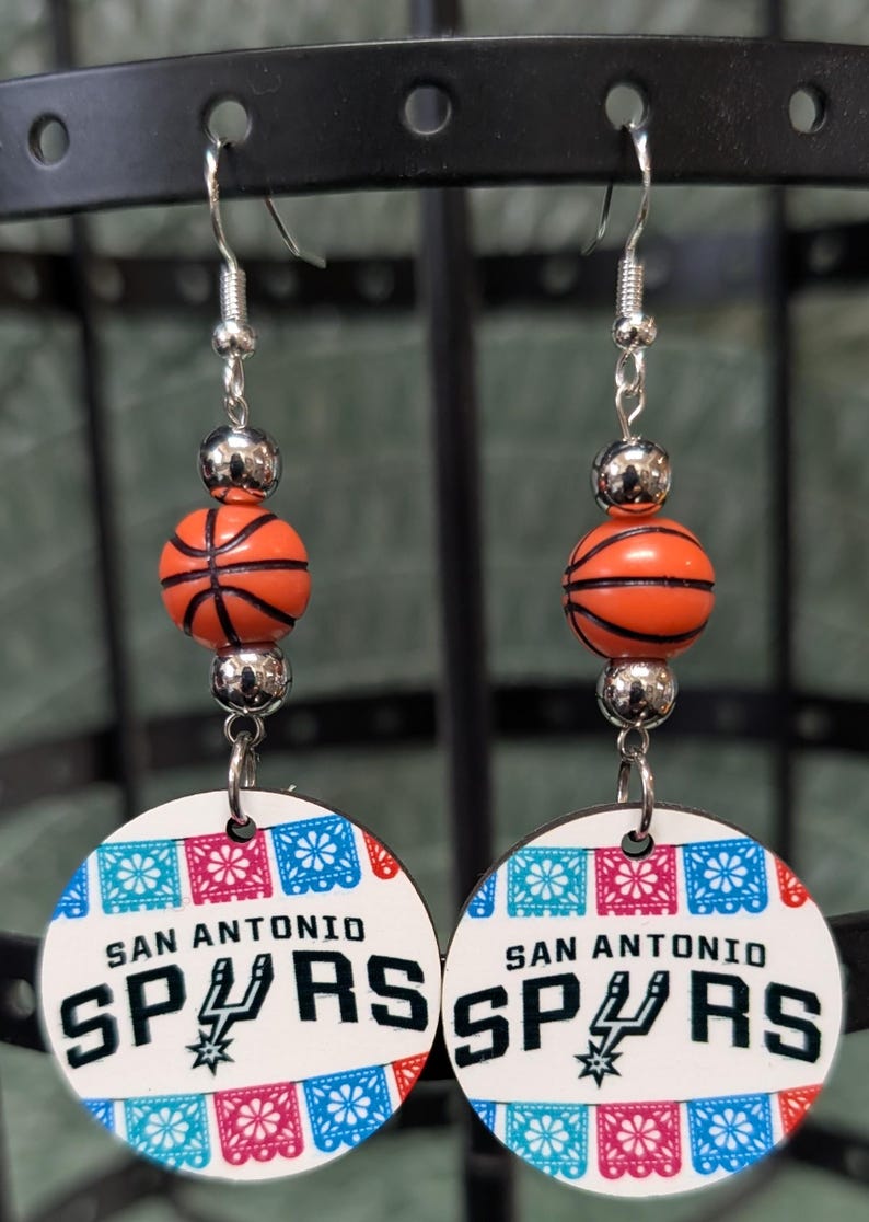 May include: A pair of dangle earrings featuring a silver-tone hook, a metallic silver bead, an orange basketball bead, and a circular white pendant. The pendant displays the text SAN ANTONIO SPURS with a team logo and colorful floral banner patterns.
