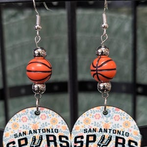 May include: A pair of dangle earrings featuring a silver-toned hook, a metallic silver bead, an orange basketball-shaped bead, and a circular pendant. The pendant displays a floral pattern with the text SAN ANTONIO SPURS and the team logo.