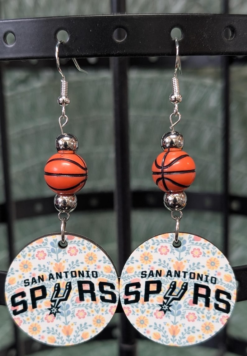 May include: A pair of dangle earrings featuring a silver-toned hook, a metallic silver bead, an orange basketball-shaped bead, and a circular pendant. The pendant displays a floral pattern with the text SAN ANTONIO SPURS and the team logo.