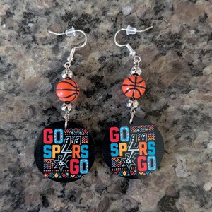 May include: A pair of dangle earrings featuring a round black pendant with the text GO SPURS GO in red, blue, and white, accented by a geometric pattern. Above the pendant is a small orange basketball bead and silver-toned metal spacers.