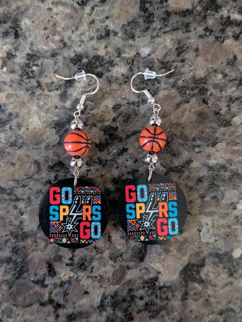 May include: A pair of dangle earrings featuring a round black pendant with the text GO SPURS GO in red, blue, and white, accented by a geometric pattern. Above the pendant is a small orange basketball bead and silver-toned metal spacers.