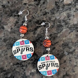 May include: A pair of dangle earrings featuring a round white pendant with the text SAN ANTONIO SPURS and a basketball team logo, accented by colorful papel picado patterns. Each earring includes a small orange basketball bead and silver hardware.