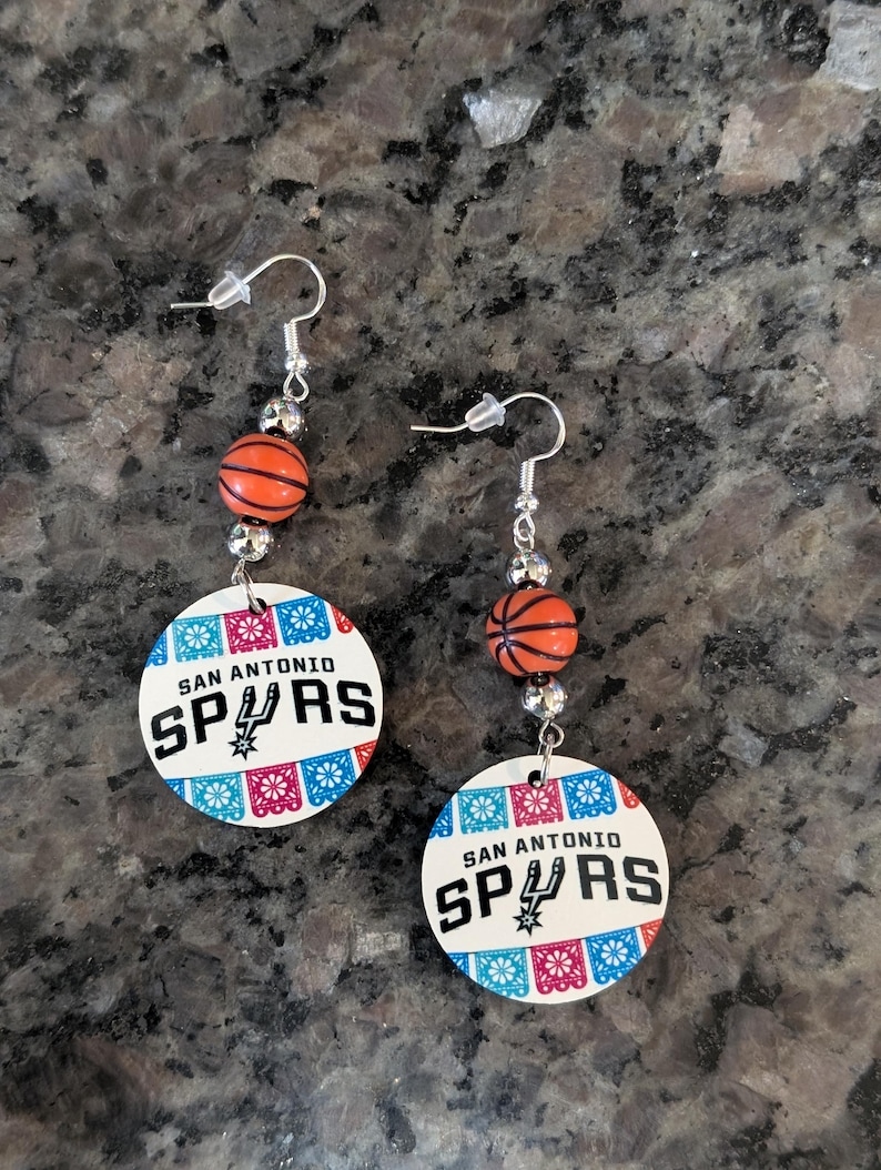 May include: A pair of dangle earrings featuring a round white pendant with the text SAN ANTONIO SPURS and a basketball team logo, accented by colorful papel picado patterns. Each earring includes a small orange basketball bead and silver hardware.
