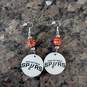 May include: A pair of dangle earrings featuring a round white disc with the black text SAN ANTONIO SPURS and a team logo. Above the disc, a small orange basketball bead is flanked by two silver-toned metallic beads on a silver-toned hook wire.