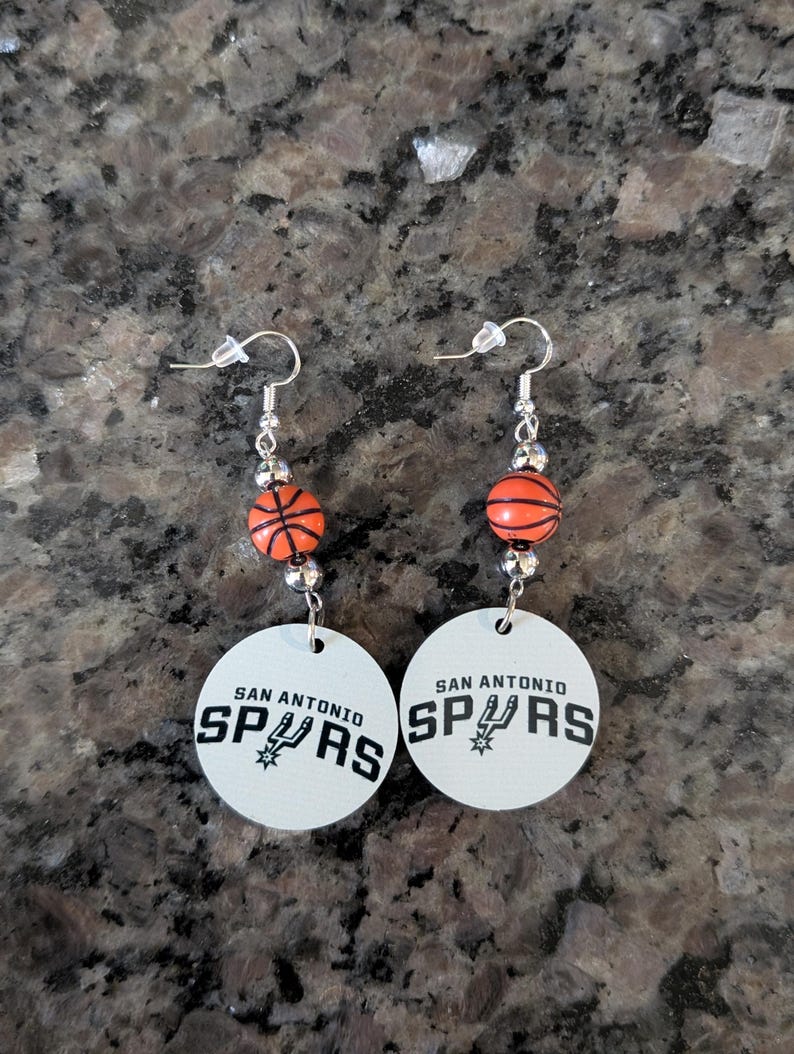 May include: A pair of dangle earrings featuring a round white disc with the black text SAN ANTONIO SPURS and a team logo. Above the disc, a small orange basketball bead is flanked by two silver-toned metallic beads on a silver-toned hook wire.