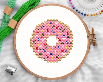 May include: A pink donut with colorful sprinkles, embroidered on white fabric in a wooden embroidery hoop.