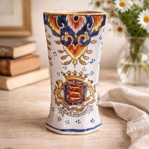 Pode incluir: Um vaso de cerâmica branco com um design detalhado e colorido. O vaso apresenta um brasão com leões, uma coroa e a palavra "Normandie". A borda superior tem uma borda dourada, e o design inclui padrões florais azuis e laranja.