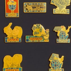 May include: A collection of vintage LUSTUCRU enamel pins. The pins feature various designs, including a chef, a bear, and cartoon figures. Each pin has the LUSTUCRU logo and is set against a dark background. The pins are in shades of yellow, blue, and red.