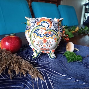 Peut inclure: Un pot de fleurs décoratif en céramique avec une base blanche et des motifs floraux colorés en orange, bleu et vert. Le pot a trois petits pieds et un bord festonné. Une pomme et du feuillage sont à proximité.