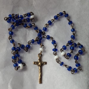 Peut inclure: Un chapelet avec des perles de verre bleues et des perles blanches en guise d'ornement, enfilées sur une chaîne métallique. Un petit crucifix doré est suspendu à la chaîne. Il s'agit d'un objet religieux.