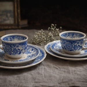 Peut inclure: Deux tasses et soucoupes assorties avec un motif floral bleu. Les tasses et les soucoupes sont blanches avec un motif floral bleu. Les tasses ont des anses. L'ensemble est sur une nappe grise.