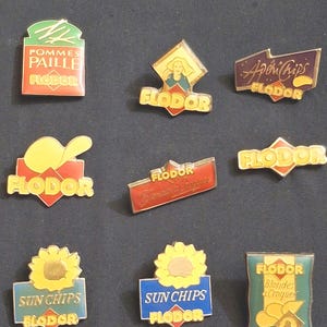 Peut inclure: Ensemble de badges émaillés vintage, principalement de la marque "FLODOR". Les motifs incluent des paquets de chips, des tournesols et des textes comme "Sun Chips" et "ApériChips".