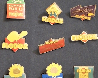 Op de afbeelding: Een verzameling vintage emaille pins, voornamelijk van het merk "FLODOR". De pins tonen verschillende ontwerpen, waaronder chipszakken, zonnebloemen en tekst zoals "Sun Chips" en "ApériChips".