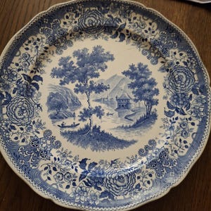 Puede incluir: Un plato decorativo redondo, blanco y azul, con borde festoneado. El plato presenta una detallada escena de paisaje azul con árboles, agua y edificios. Patrones y diseños florales rodean la imagen central. El plato está sobre una superficie de madera.