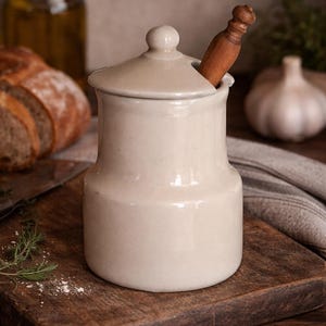 Puede incluir: Un tarro de cerámica color crema con tapa y cuchara de madera. El tarro está sobre una tabla de cortar de madera con pan, ajo y hierbas en el fondo. El tarro mide aproximadamente 15 cm de alto.
