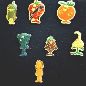 May include: A collection of colorful enamel pins featuring cartoon fruit and vegetable characters. The pins include a strawberry, tomato, pineapple, and other whimsical designs with gold-colored outlines. The pins are set against a dark background.