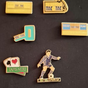 May include: A collection of gold-toned enamel pins. The pins feature text and graphics, including playing cards, a soccer player, and the words "TAC TAC" and "Tapis Vert". The pins are likely collectibles or promotional items.