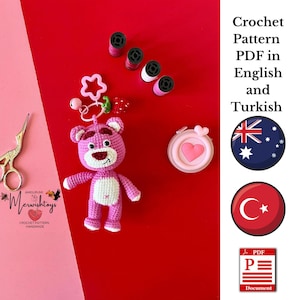 May include: Pink crocheted teddy bear amigurumi toy with a white belly and a pink heart on its chest. The bear is attached to a pink and green strawberry charm and a pink star charm. The toy is on a red background with yarn and a pair of scissors. The image also includes text that says "Amigurumi Crochet Pattern Handmade" and "Merwishtoys".