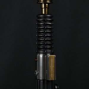 Puede incluir: Una réplica de sable de luz plateado y negro con una parte superior de oro y cobre. El sable de luz tiene un agarre negro texturizado y un soporte de hoja plateado.