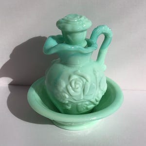 K&ouml;nnte beinhalten: Ein Vintage-Waschtisch-Set aus hellgr&uuml;nem Glas. Das Set umfasst einen kleinen Krug mit Rosenmuster und ein passendes Becken. Der Krug hat einen Griff und einen dekorativen Deckel. Das Set besteht aus undurchsichtigem Glas.