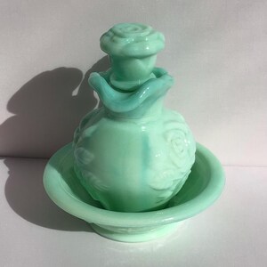 K&ouml;nnte beinhalten: Ein Vintage-Parf&uuml;mflaschen-Set aus hellgr&uuml;nem Glas. Die Flasche hat einen rosenf&ouml;rmigen Verschluss und ist mit Rosenmotiven verziert. Sie steht in einer passenden flachen Schale. Das Set hat eine weiche, pastellgr&uuml;ne Farbe.