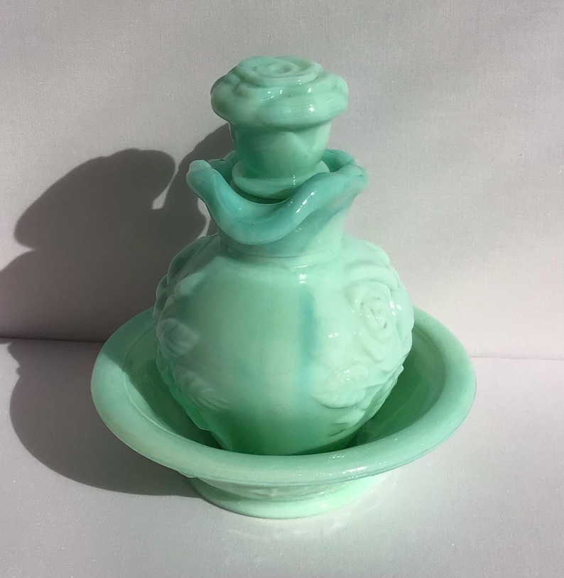 K&ouml;nnte beinhalten: Ein Vintage-Parf&uuml;mflaschen-Set aus hellgr&uuml;nem Glas. Die Flasche hat einen rosenf&ouml;rmigen Verschluss und ist mit Rosenmotiven verziert. Sie steht in einer passenden flachen Schale. Das Set hat eine weiche, pastellgr&uuml;ne Farbe.