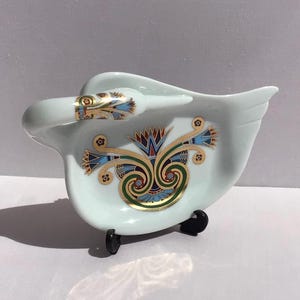 May include: A pale blue ceramic swan-shaped dish featuring a decorative gold, blue, and green floral pattern on its body and neck. The swan has a curved neck and wing-like edges, resting on a small black plastic stand for display purposes.