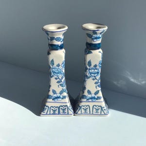 May include: A pair of ceramic candle holders featuring a white base with blue floral patterns and a geometric border design at the bottom. Each holder has a flared top and a square base, showcasing a consistent hand-painted style throughout.