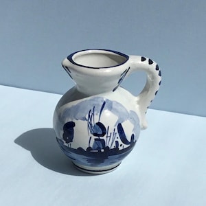 May include: A small, white ceramic jug with a blue and white Delftware design. The jug has a rounded body and a short, curved spout. The handle is attached to the side of the jug and is also decorated with blue and white Delftware.