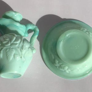 K&ouml;nnte beinhalten: Ein Vintage-Sahne- und Untertassen-Set aus hellgr&uuml;nem Glas. Der Sahnebereiter hat ein Rosendesign und einen Griff. Die Untertasse hat ein passendes Blumenmuster. Das Set stammt wahrscheinlich aus der Mitte des 20. Jahrhunderts.