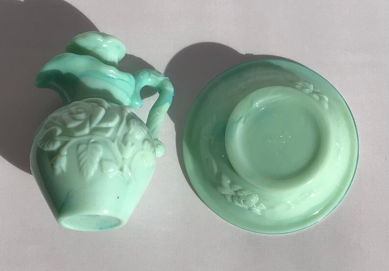 K&ouml;nnte beinhalten: Ein Vintage-Sahne- und Untertassen-Set aus hellgr&uuml;nem Glas. Der Sahnebereiter hat ein Rosendesign und einen Griff. Die Untertasse hat ein passendes Blumenmuster. Das Set stammt wahrscheinlich aus der Mitte des 20. Jahrhunderts.