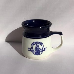 May include: A ceramic mug with a white body and a navy blue rim and interior. The mug features a handle and a blue emblem with the text "CELEBRATING 1988". The mug is designed with a classic, vintage aesthetic.