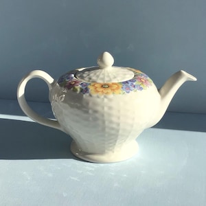 May include: A white ceramic teapot featuring a textured basket-weave pattern on the body and lid. The upper rim of the teapot and the edge of the lid are decorated with a colourful floral band of yellow, purple, and blue flowers.
