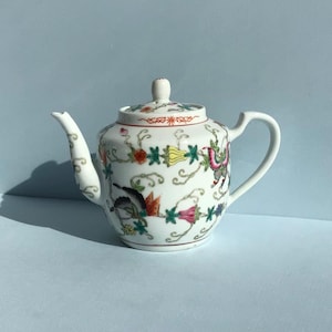 May include: A white porcelain teapot featuring a colourful hand-painted pattern of butterflies, stylised flowers, and swirling green vines. The teapot includes a matching lid with a small knob, a curved spout, and a handle, all decorated with fine lines.